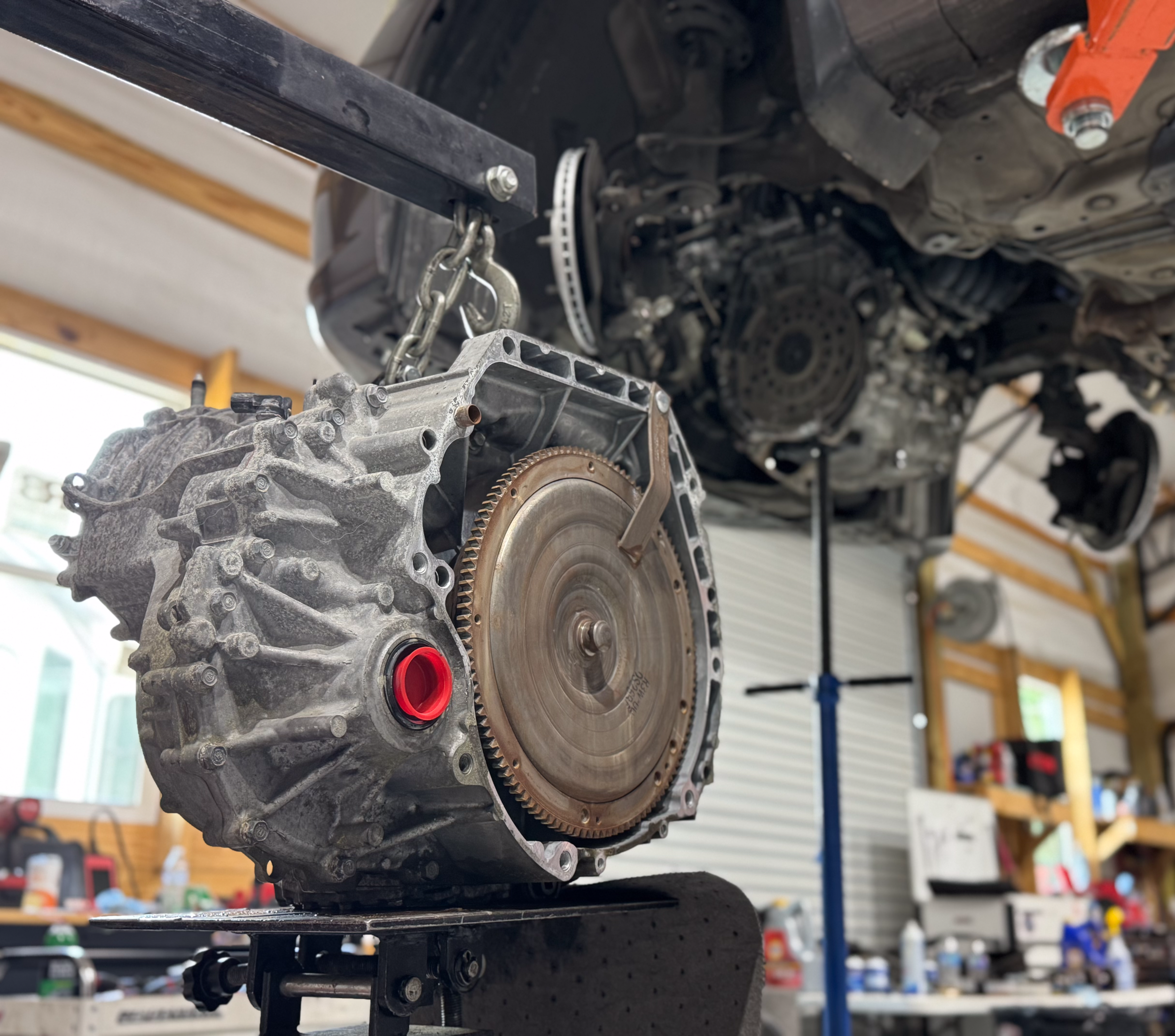 A picture of a transmission in the foreground sitting on a transmission jack waiting to be installed into the vehicle in the background